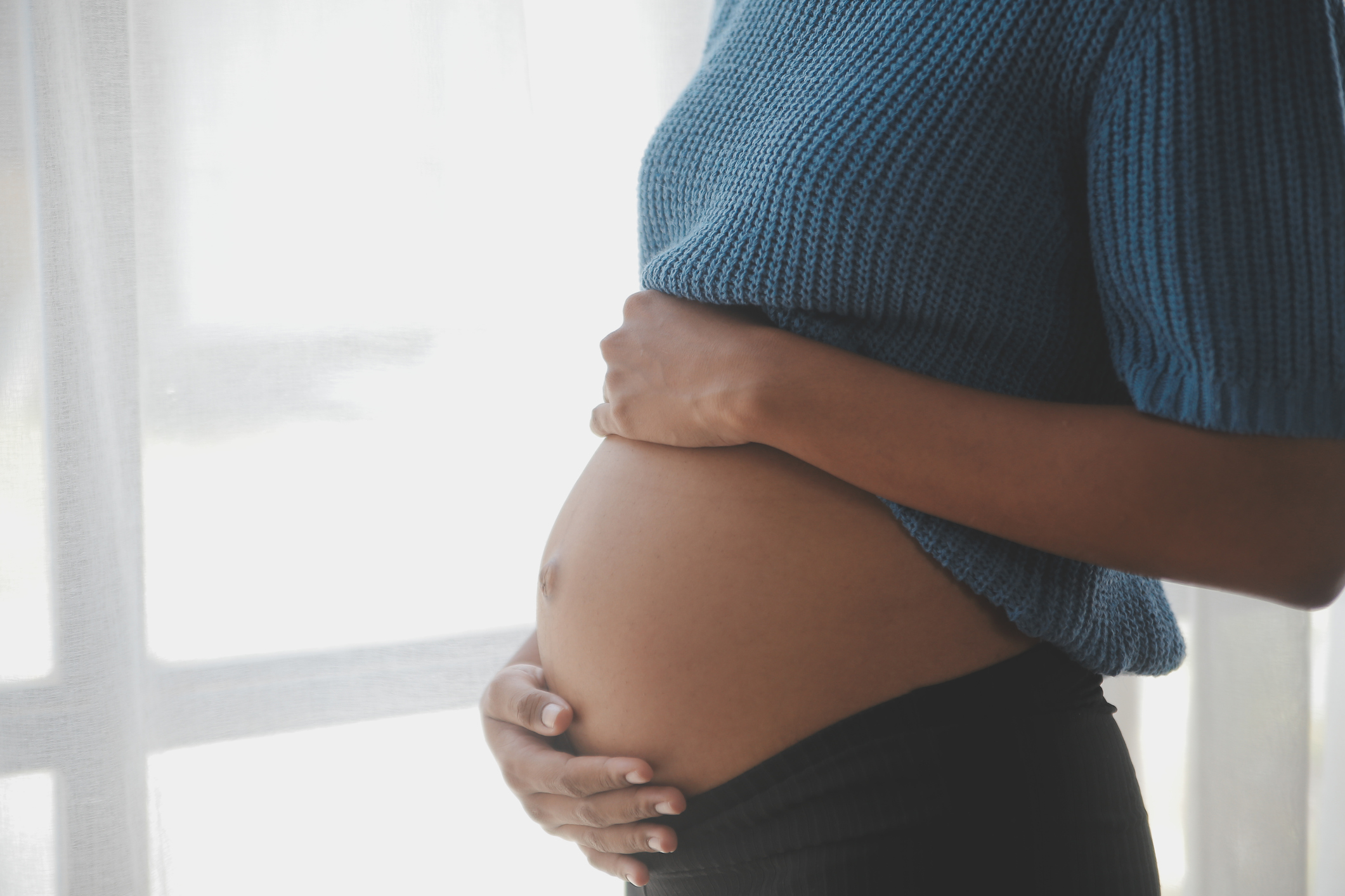 Schwangere Frau, die ihren Bauch hält. Sie trägt einen blauen Pullover und schwarze Hose. Helle Fenster im Hintergrund.